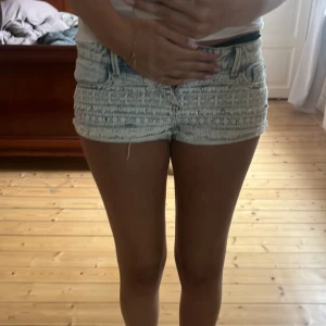 Ljusblå jeansshorts med spetsdetaljer - Supersnygga ljusblå jeansshorts från Italy med slitna kanter och spetsdetaljer framtill. Shortsen har normal passform, klassiska fickor och små nitar på spetsen för extra edge. Perfekta till sommaren och ger en cool, feminin vibe. Storleken är 32. 