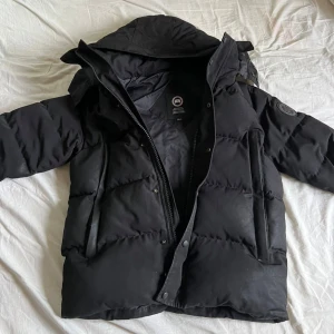Canada Goose jacka  - Säljer en svart jacka från Canada Goose med huva och diskret logga på ärmen. Jackan är vadderad, har dragkedja och knappar framtill. Säljer åt min bror då den blivit för liten på honom. Den är äkta och är i fint skick! Ny pris cirka 10-15 tusen.