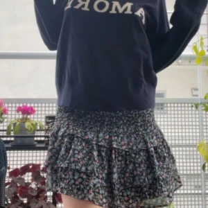 Blommig volangkjol i svart och rosa,blå och grön  - Supersöt kort kjol med blommigt mönster i svart, rosa,blått grönt. Kjolen har volanger och en smockad midja. 