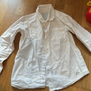 Vit klassisk skjorta i bomull - Vit skjorta i klassisk modell med lång ärm, bröstficka och knappar framtill. Skjortan är tillverkad i mjuk bomull och har en enkel, stilren look som passar till det mesta. Perfekt för dig som gillar en clean och tidlös stil.