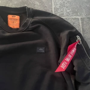 Svart sweatshirt från Alpha Industries - Oanvänd Svart sweatshirt från Alpha Industries med klassisk rund hals och ribbade muddar. Tröjan har en ikonisk röd tag på ärmen med dragkedja och en liten logotyp på bröstet. Perfekt för dig som gillar streetwear och snygga detaljer.