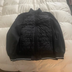 Svart quiltad bomberjacka - Svart bomberjacka med quiltad front och släta ärmar. Jackan har dragkedja framtill och ribbade muddar med vita ränder vid ärmslut och nederkant. Perfekt för dig som vill ha en stilren och sportig look. Storlek S, men kan passa M.