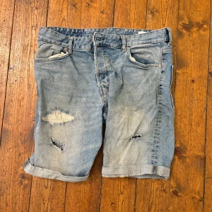 Slim fit denimshorts ljusblå - Slim fit jeansshorts i ljusblå denim med slitna detaljer och lagade hål framtill. Klassisk femficksmodell, uppvikta benslut och midwaist. Perfekt för dig som gillar en avslappnad och edgy stil. 