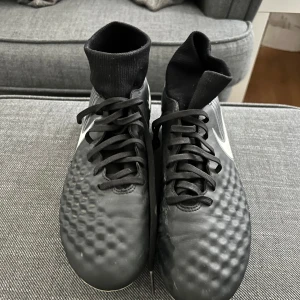 Svarta Nike fotbollsskor med socka - Snygga svarta Nike fotbollsskor med högt skaft i strumpmodell och stora vita swoosh-loggor på sidorna. Ovandelen har en texturerad yta för bättre bollkontroll och snörning framtill. Sulan är vit med gröna dobbar, perfekt för gräsplan.