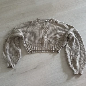 Beige croppad stickad tröja NA-KD - Superfin beige croppad stickad tröja från NA-KD med breda ärmar och ribbade muddar. Snygg kabelstickad detalj framtill och rund halsringning. Perfekt till höst- och vinterlooken, passar till både jeans och kjol.