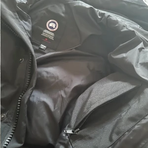 Svart Canada Goose - Säljer en svart dunjacka från Canada Goose med huva och klassisk logga på ärmen. Jackan har dragkedja, knappar och flera fickor, perfekt för kalla dagar. Fodrad och riktigt varm, passar dig som vill ha en stilren och funktionell vinterjacka.