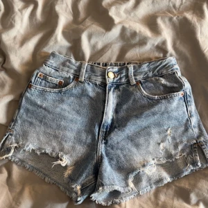 Blå jeansshorts med fransig kant från Zara - Snygga blå jeansshorts från Zara med fransig kant och slitna detaljer. Klassisk femficksmodell, knapp och dragkedja framtill samt elastisk midja bak. Perfekta för varma dagar och har en cool, avslappnad vibe.