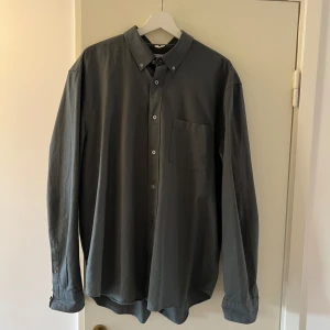 Mörkgrå/mörkgrön skjorta Regular Fit strl 54 - Snygg mörkgrå/mörkgrön skjorta i regular fit med klassisk krage och knappstängning framtill. Skjortan har en bröstficka och långa ärmar med manschett. Perfekt för dig som gillar en stilren och enkel look. Köpt på arket för ca 3 månader sedan och använd ca 5 tillfällen. Köpt för 800kr. Storleken är typ en L-XL. Se bild 3 för uppfattning av färgen. Den ser mörk grå ut i vissa ljus och mörk grön ut i vissa ljus