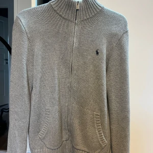 Grå stickad kofta från Polo Ralph Lauren - Snygg grå stickad kofta från Polo Ralph Lauren med dragkedja framtill, ribbad hög krage och två fickor. Klassisk logga broderad på bröstet och coola ränder i vinrött och marinblått i nacken. Perfekt för lager-på-lager och chill stil.