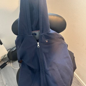 Marinblå hoodie från GANT med dragkedja - Snygg marinblå hoodie från GANT med hel dragkedja och huva. Klassisk design med två stora fickor framtill och diskret GANT-logga på bröstet. Perfekt för chill dagar och enkel att matcha med jeans eller joggers.