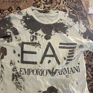 EA7 Emporio Armani t-shirt med tryck - Snygg EA7 Emporio Armani t-shirt i ljusgrått med mörkgrått splattermönster och stort logotryck på bröstet. Klassisk rund hals och korta ärmar. Tillverkad i mjuk bomull som känns skön mot huden. Perfekt för dig som gillar streetwear och vill sticka ut. Pris går såklart diskuteras