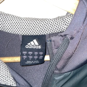 Svart vindjacka från Adidas - Svart vindjacka från Adidas med huva och hel dragkedja. Jackan har sportig design, meshfoder och snörning i huvan. Perfekt för dig som gillar en clean och funktionell look. Adidas-logga på bröstet och praktiska fickor på sidorna.