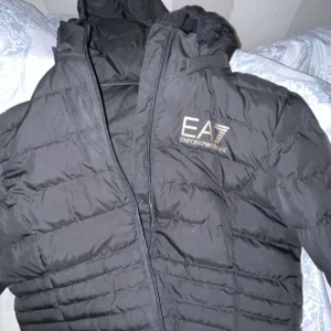 Svart dunjacka från EA7 Emporio Armani - Säljer en svart dunjacka från EA7 Emporio Armani med diskret logga på bröstet. Jackan har en klassisk pufferstil, dragkedja framtill och hög krage. Perfekt för kalla dagar när du vill hålla stilen och samtidigt vara varm.