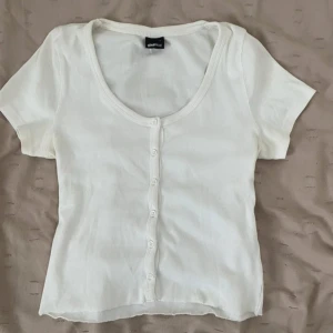 Vit ribbad topp med knappar Gina Tricot - Superfin vit ribbad topp från Gina Tricot med korta ärmar och söt knappdetalj framtill. Modellen har en rundad urringning och är figurnära i passformen. Perfekt att styla med jeans eller kjol för en fräsch look.