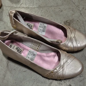 Ballerinaskor - Snygga ballerinaskor från skopunkten i beige metallic med låg kilklack. Skorna har rund tå, dekorativa remmar framtill och små guldfärgade nitar. Insidan är fodrad med ljusrosa tyg för extra stilpoäng. Perfekta för dig som gillar en feminin look med lite höjd.