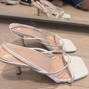 Vita sandaler med klack och smala remmar - Snygga vita sandaler med smala remmar och fyrkantig tå. Klacken är smal och ger en elegant look. Ytan har ett diskret texturmönster som ger extra stilpoäng. Perfekta för dig som gillar minimalistisk design och vill ha något fräscht till sommaren.