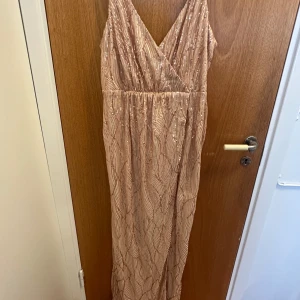 Champagnefärgad paljettklänning med slits - Säljer en lång, champagnefärgad paljettklänning från Henriette med v-ringning och omlottliv. Klänningen har en snygg slits framtill och glittrigt mönster som ger en lyxig känsla. Perfekt för dig som vill sticka ut och glänsa på festen!