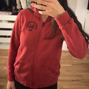 Röd kofta från Peak Performance - Säljer en röd hoodie från Peak Performance med svart broderad logga och siffran 86 på bröstet. Tröjan har dragkedja, huva och två fickor framtill. Mjuk insida och ribbade muddar vid ärmslut och nederkant. Perfekt för chill dagar eller när du vill vara bekväm.