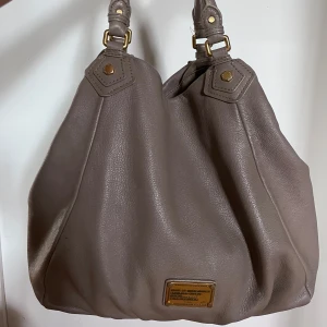 Beige axelväska från Marc by Marc Jacobs - Snygg beige axelväska i mjukt skinn med guldfärgade metalldetaljer och justerbara remmar. Insidan har svartvitt tryck med logotypen. Handtagen är lite slitna annars väldigt bra skick.
