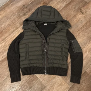 Moncler dunjacka med stickade ärmar - Snygg mörkgrön dunjacka från Moncler med stickade svarta ärmar och huva. Jackan har quiltad front, dragkedja, ribbade muddar och en liten ficka på ärmen. Perfekt för dig som vill ha en sportig och trendig look under höst och vinter. Scan funkar och priset är inte hugget i sten