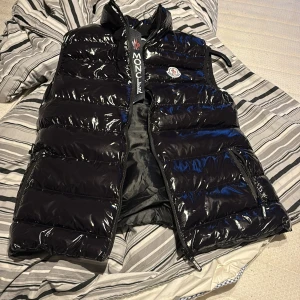 Moncler väst - Lyxig Moncler-väst – Storlek L  Snygg och stilren Moncler-väst i blank svart finish. Perfekt för dig som vill höja din outfit med ett exklusivt plagg som både är bekvämt och trendigt. Västen är lätt, varm och fungerar lika bra till vardags som vid finare tillfällen.  ✔️ Storlek: L ✔️ Färg: Svart (glansig) ✔️ Klassisk Moncler-design med logga på bröstet ✔️ Passar både höst, vinter och vår  Nypris på Moncler-västar ligger betydligt högre – här får du chansen att göra ett riktigt kap för endast 1600kr.