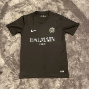 Svart PSG x Balmain fotbollströja Nike - Svart fotbollströja från Nike i samarbete med PSG och Balmain. Tröjan har korta ärmar, rund hals och tryck med Balmain Paris samt PSG-logga på bröstet. Materialet är lätt och andas, perfekt för träning eller match. Snygg och stilren design med vit logotyp.