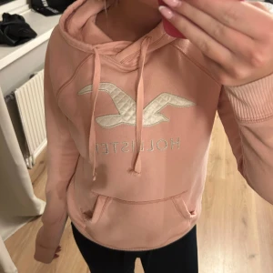 Ljusrosa hoodie från Hollister - Säljer en ljusrosa hoodie från Hollister med stor broderad logga framtill. Tröjan har huva med snörning, magficka och långa ärmar med ribbade muddar. 