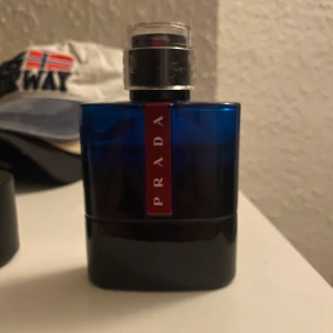 Prada Luna Rossa ocean edt - Snygg parfymflaska från Prada Luna Rossa-serien i en djupblå färg med svart botten och en röd vertikal etikett. Flaskan har en modern, stilren design och är tillverkad i glas med en metallisk spraykork. Perfekt för dig som gillar exklusiva dofter. Ungefär 75/80ml kvar av 100 som ni kan se😁