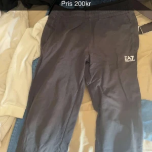 Svarta mjukisbyxor från EA7 - Svarta mjukisbyxor från EA7 Emporio Armani med elastisk midja och logga tryckt på vänster lår. Perfekta för chill eller träning, med en enkel och clean look. Sköna och lätta att matcha med hoodies eller t-shirts.