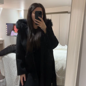 Svart pälskappa - Säljer en svart kappa från femenina of sweden med stor fluffig päls runt huvan. Kappan har ett bälte i midjan och guldigt märke på ärmen. Perfekt för kalla dagar när du vill vara både varm och snygg. Har används endast 5 gånger 