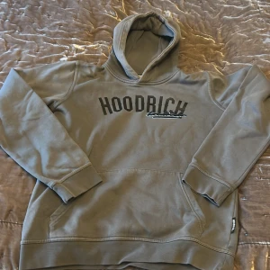 Grå hoodie från Hoodrich med tryck - Säljer en grå hoodie från Hoodrich med svart tryck på bröstet och klassisk känguruficka. Tröjan har huva och ribbade muddar vid ärmslut och nederkant. Perfekt för dig som gillar streetwear och vill ha en stilren look.