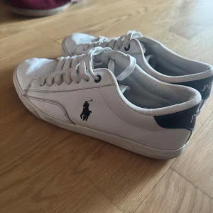Vita sneakers från Polo Ralph Lauren - Klassiska vita sneakers från Polo Ralph Lauren med svart logga på sidan och svart detalj med POLO-text på hälen. Skorna har snörning och rund tå, tillverkade i skinn och textil. Perfekta för en clean och stilren look.