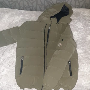Olivgrön Moncler dunjacka med huva - Säljer en olivgrön dunjacka som gör att du står ut från alla andra och har din egen stil, dunjackan är från Moncler i storlek M. Jackan har huva, svarta dragkedjor på fickor och framtill, samt Moncler-logga på ärmen. Fodret är svart och jackan har justerbar dragsko nertill. Perfekt för kalla dagar.