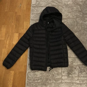 Svart pufferjacka från Pier One - Svart pufferjacka från Pier One med huva och horisontella sömmar. Jackan har dragkedja framtill och är lätt vadderad, perfekt för kyliga dagar. Enkel och stilren design som funkar till det mesta. Materialet är syntet och jackan har lång ärm.