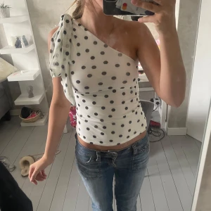 Prickig one shoulder topp från H&M Divided - (Aldrig använt)Snygg vit one shoulder topp från H&M Divided med svarta prickar och stor knytrosett på axeln. Toppens passform är tight och den är tillverkad i ett stretchigt polyestertyg. Perfekt för dig som vill sticka ut med en cool och trendig look.