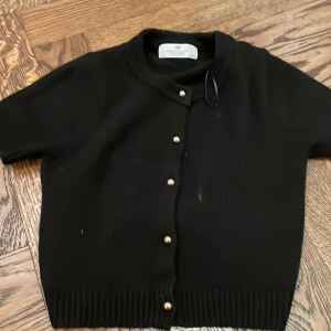 Svart kofta med pärlknappar från H&M - Kortärmad svart kofta från H&M med  pärlknappar framtill. Koftan är stickad med ribbade muddar vid ärmslut och nederkant. Storlek 134/140