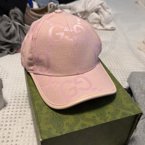 Rosa Gucci keps - Snygg rosa keps från Gucci med ikoniskt GG-mönster i ljuslila över hela kepsen. Kepsen har böjd skärm och justerbar rem baktill. Tillverkad i tålig bomullscanvas med exklusiv känsla. Perfekt för dig som vill sticka ut med en lyxig accessoar. Låda ingår inte