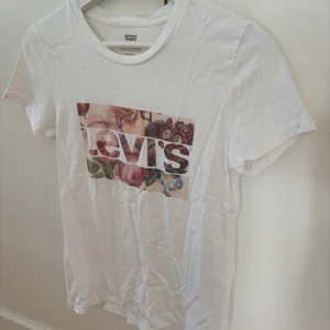 Vit Levi's t-shirt med blommigt tryck - Vit t-shirt från Levi's med klassiskt logotryck över bröstet, där bakgrunden är fylld med färgglada blommor. Modellen är kortärmad och har rund hals. Perfekt för dig som gillar stilrena plagg med en twist.