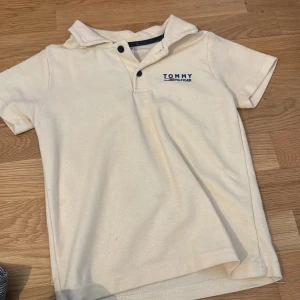 Beige piké från Tommy Hilfiger - Snygg beige pikétröja från Tommy Hilfiger med klassisk krage och två knappar framtill. T-shirten har korta ärmar och logga broderad på bröstet och ärmen. Perfekt för en clean och stilren look.
