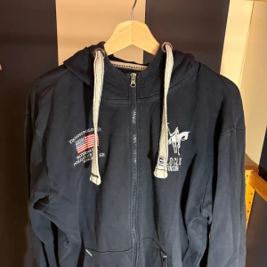 Svart hoodie från Vinson Polo Club - Svart hoodie från Vinson Polo Club med broderad logga och amerikansk flagga på bröstet. Tröjan har dragkedja, stora fickor framtill, huva med breda snören och är tillverkad i mjuk bomull. Perfekt för en chill och sportig stil.