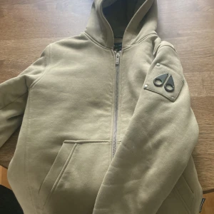 Beige hoodie från Moose Knuckles XL - Snygg beige hoodie från Moose Knuckles i storlek XL barn passar från 170 till -175T nästan helt ny tröjan har dragkedja framtill, huva och en ikonisk logga på ärmen. Mjuk insida och praktisk ficka framtill. Perfekt för dig som gillar streetwear och vill ha något stilrent men ändå unikt.