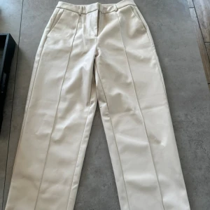 Beiga kostymbyxor från ONLY - Stilrena beiga kostymbyxor från ONLY med pressveck och raka ben. Byxorna har hällor för bälte, sidfickor och två bakfickor. Snyggt midjehöga och tillverkade i ett slätt material som ger en clean look.