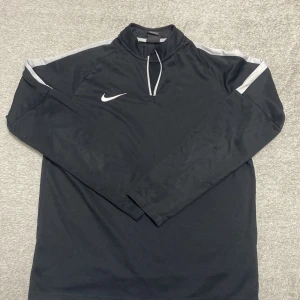 Svart Nike Dri-Fit half zip tröja XL - Svart långärmad Nike Dri-Fit tröja med grå detaljer på axlarna och vit Nike-logga på bröstet. Tröjan har half zip och är tillverkad i ett lätt, syntetiskt material som andas. Perfekt för träning eller chill dagar. Zipen är även sönder 