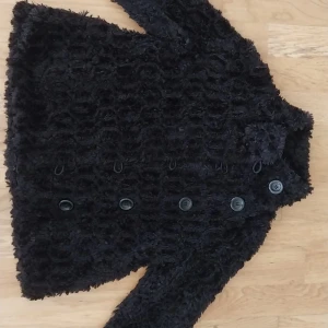 Svart teddyjacka från Lindex - Mysig svart teddyjacka från Lindex med fluffig struktur och dubbla rader knappar framtill. Jackan har lång ärm och är perfekt för kyligare dagar. Snygg och trendig look som passar till många outfits.