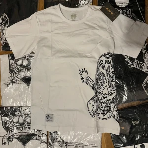 Vit t-shirt Dirty Rebel Wear med tryck - Snygg vit t-shirt från Dirty Rebel Wear med coolt svart grafiskt tryck på sidan. Rund hals och korta ärmar, tillverkad i mjuk bomull. Märkespatch nertill och unik design som sticker ut. Perfekt för dig som gillar streetstyle och edgy prints.