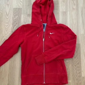 Röd Nike hoodie med dragkedja - Röd hoodie från Nike med vit logga på bröstet. Tröjan har huva med snörning, hel dragkedja och två fickor framtill. Riktigt snygg och varm. Perfekt för varje dag men även träning. Mycket bra skick knappast använt