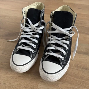 Svarta Converse Chuck Taylor High - Säljer ett par klassiska Converse Chuck Taylor All Star high tops i svart canvas med vita gummisulor och vita snören. Skorna har rund tå, platt sula och den ikoniska loggan på sidan. Perfekt för dig som gillar retrostil och vill ha ett par tidlösa sneakers.