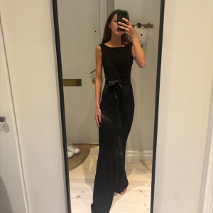 Svart långklänning med sidenband - Svart långklänning med sidenband i midjan från Carolina Gynnings kollektion med bubbleroom.  Använd enbart 1 gång. Observera dock att den ena hållaren för midjesnöret har lossnat. Midjebandet kan dock användas som vanligt ändå. Perfekt för dig som vill ha en stilren och tidlös look. ❤️
