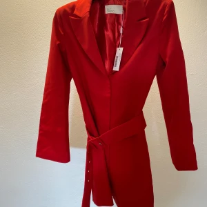 Röd kavajklänning från NLY Trend - Snygg röd kavajklänning från NLY Trend med markerad krage, långa ärmar tillhörande bälte i midjan. Klänningen har en stilren, figurnära passform och är tillverkad i ett glansigt polyestermaterial som ger en lyxig känsla. Perfekt för dig som vill sticka ut.