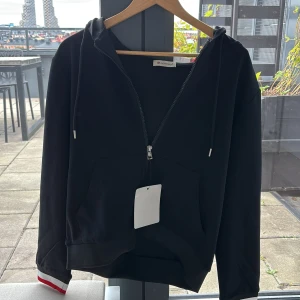 Moncler zip up hoodie - Svart hoodie från Moncler med hel dragkedja framtill, huva med snörning och klassisk känguruficka. Ärmarna har en snygg detalj med röd och vit rand vid mudden. Tillverkad i mjuk bomull och prydd med Moncler-logga på ärmen.